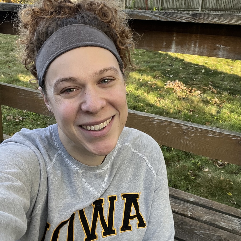 A young woman in a grey sweatshirt with the University of Iowa logo on it, smiling at the camera. She has blue eyes and curly hair, pulled into a short ponytail.